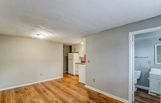 A room with wooden floors and a white ceiling with a kitchen area visible through the door.