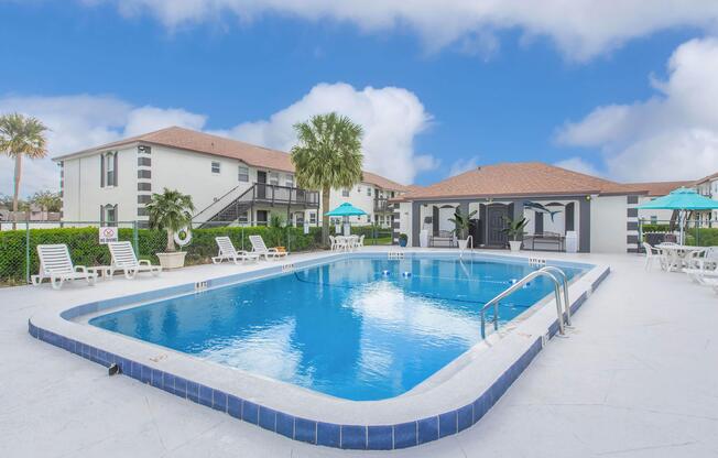 A clear blue swimming pool surrounded by lounge chairs and tropical greenery. In the background, a two-story building with a red tiled roof and umbrella-covered seating areas. The sky is bright and cloudy, creating a serene and inviting atmosphere.