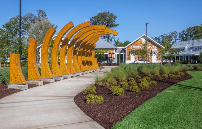 A curved wooden structure forms a visually striking archway in a landscaped park. A pathway winds through green lawns and plants, leading to a building with a sign that reads "Port Cavava." The scene is set under a clear blue sky, creating a welcoming atmosphere.