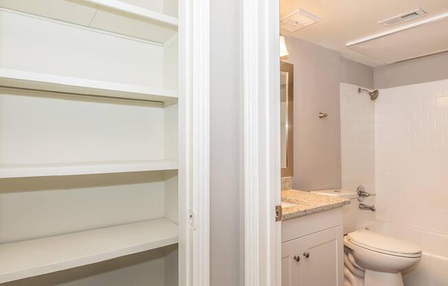 A modern bathroom featuring a white toilet, granite countertop, and a shower/tub combination. Adjacent to the bathroom is a shelf unit with empty white shelves, showcasing a clean and minimalistic design. The walls are painted in a soft gray color, enhancing the overall brightness of the space.