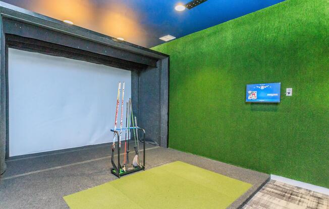Indoor golf simulator room featuring a green artificial turf area, a white projection screen, and a golf club rack with several clubs. The walls are painted dark blue and bright green, creating a vibrant atmosphere for practice.