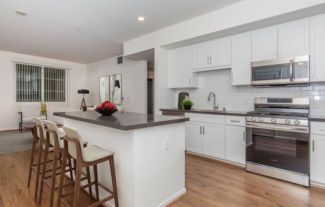 Modern kitchen with white cabinetry, stainless steel appliances, and a gray countertop. Bar stools are arranged at the breakfast bar. In the background, a dining area is visible with a small table and decorative items. Light wood flooring enhances the bright, spacious feel of the living area.