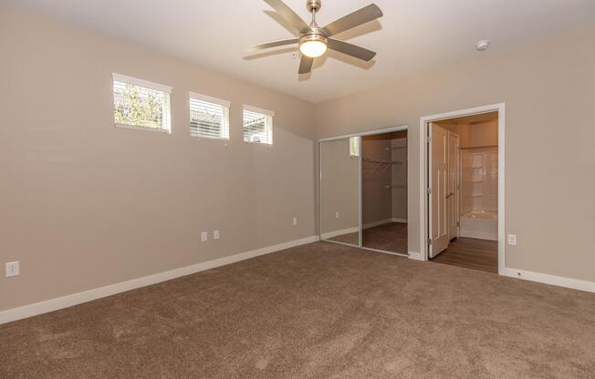 A spacious, unfurnished bedroom featuring light beige walls, a ceiling fan, and three small windows that allow natural light. On one side, there is a mirrored closet and a doorway leading to a bathroom area with white tiles. The floor is covered in soft, light brown carpet.