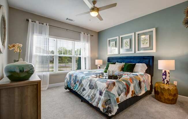 Bedroom. Big bed with two nightstands, light carpet, dresser, big windows, and ceiling fan at Ascent at Mallard Creek Charlotte, NC