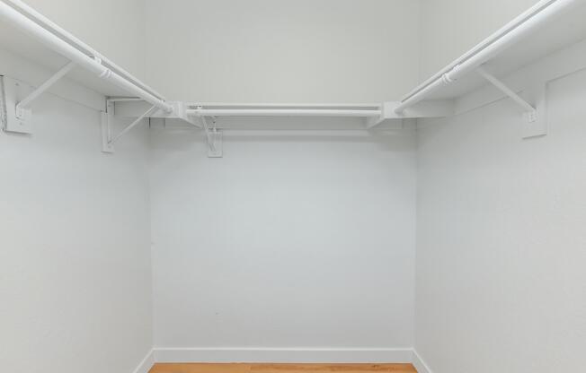 Empty closet with white walls and a wooden floor, featuring two white metal hanging rods on either side for clothes storage.