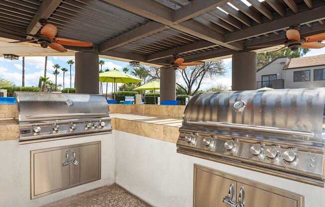 A stainless steel grill is on a patio with a ceiling fan.