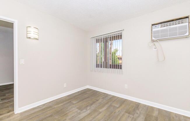 A bright, minimally furnished room featuring light-colored walls and wooden flooring. A window with vertical blinds lets in natural light, while a wall-mounted air conditioning unit is visible. The room has a modern light fixture and an empty space, suggesting it is ready for furniture or personal decoration.
