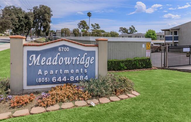 Sign for Meadowridge Apartments located at 6700 with the phone number (805) 644-8484, surrounded by landscaped greenery and flowers. The background features an entry gate and residential buildings under a clear blue sky.