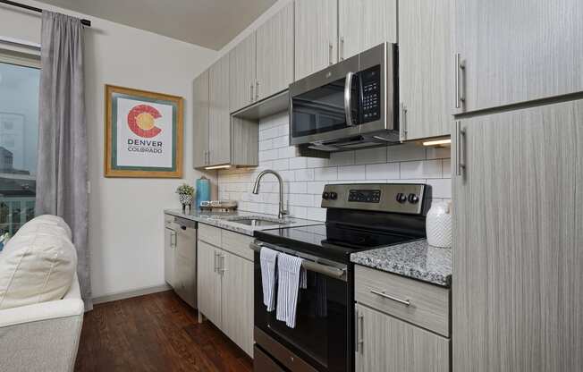 Half kitchen with wood textured gray cabinets, granite countertop and stainless steel appliances, open to living room