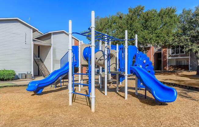 Colorful playground equipment with blue slides and climbing structure on a sandy ground, surrounded by residential buildings and trees under a clear blue sky.