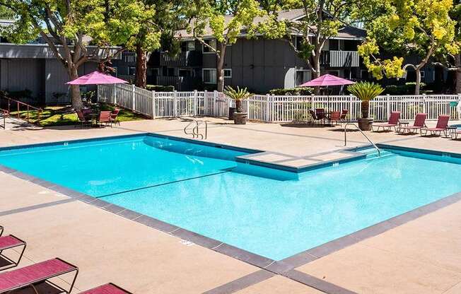 A large swimming pool surrounded by a white fence and red lounge chairs.