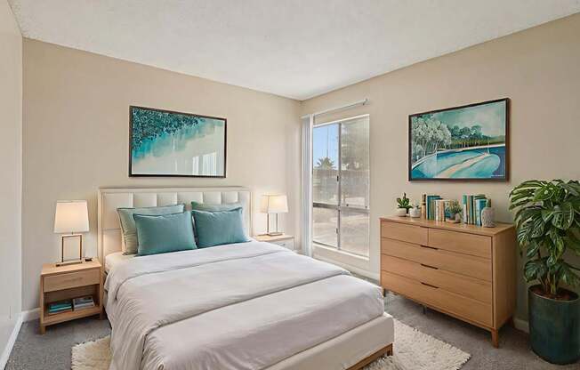 A bedroom with a large bed, a nightstand with a lamp, a dresser with a plant, and two framed pictures on the wall.