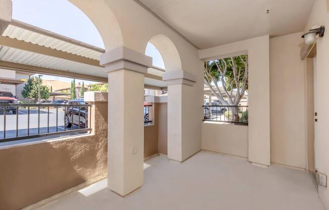A spacious patio area with arched entrances, featuring a clean concrete floor and light-colored walls. Outdoor view includes trees and parked cars behind a wrought-iron fence, providing a bright and airy atmosphere.