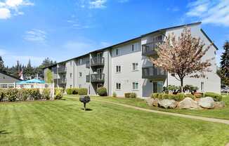 a grassy area with a tree in front of an apartment building at Pacific Park Apartment Homes, Edmonds, WA