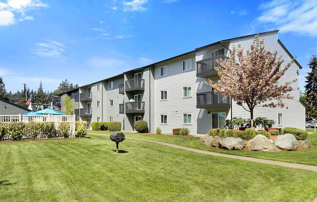 a grassy area with a tree in front of an apartment building at Pacific Park Apartment Homes, Edmonds, WA