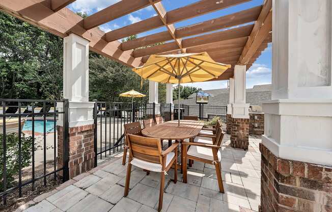 A wooden table and chairs are set up on a patio with a yellow umbrella.