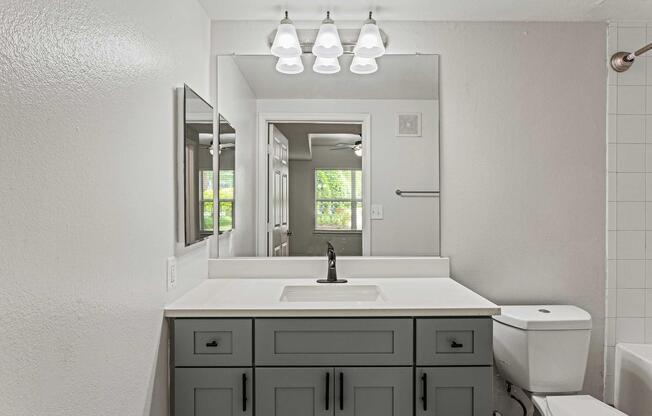 A modern bathroom featuring a gray vanity with a white sink, a large mirror above, and three pendant lights. The space includes a toilet and a tiled shower area, with a window allowing natural light to enter, highlighting a clean and contemporary design.