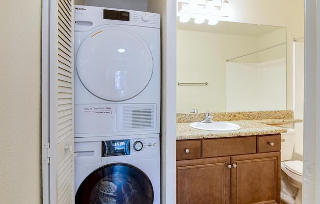A compact laundry setup featuring a stacked washer and dryer unit next to a small bathroom. The bathroom includes a sink with a granite countertop, a mirror, and a shower area with glass doors. The walls are painted in a light color, creating a bright and airy feel.