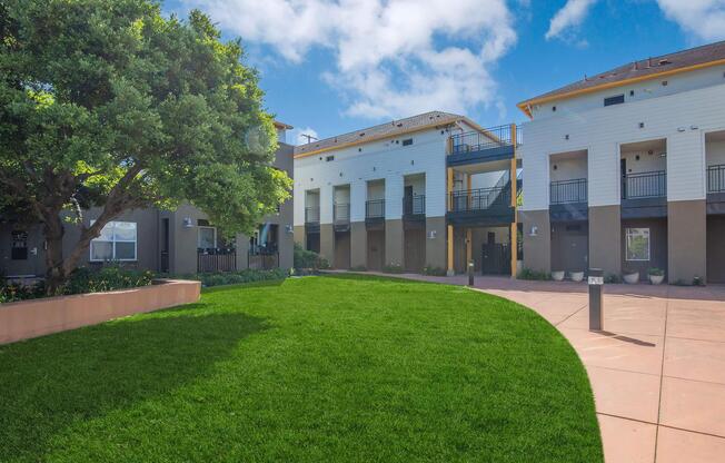A well-maintained outdoor area of an apartment complex featuring lush green grass, colorful landscaping, and modern buildings with balconies under a blue sky with a few clouds. The design includes walkways and planters, creating an inviting atmosphere for residents.