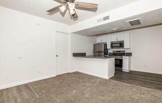 Empty living area with a ceiling fan, featuring light-colored walls and carpeted flooring. An adjoining kitchen area with modern appliances, including a microwave and stove, and a dark countertop. A closed door leads to another room, while the space appears well-lit and clean.