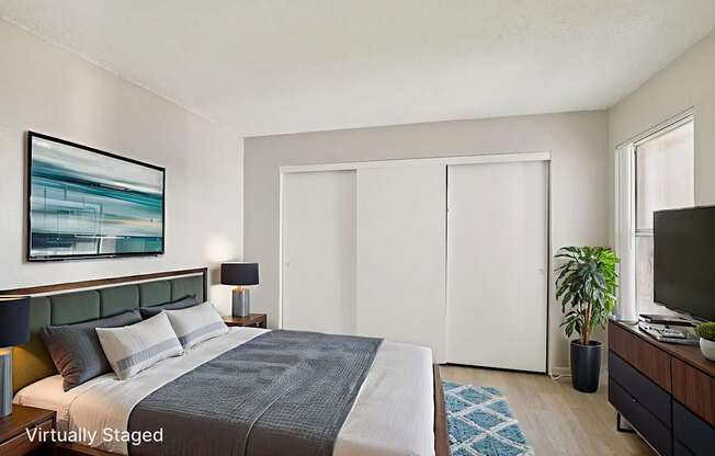 A bedroom with a bed, a painting, a lamp, a plant, and a TV.