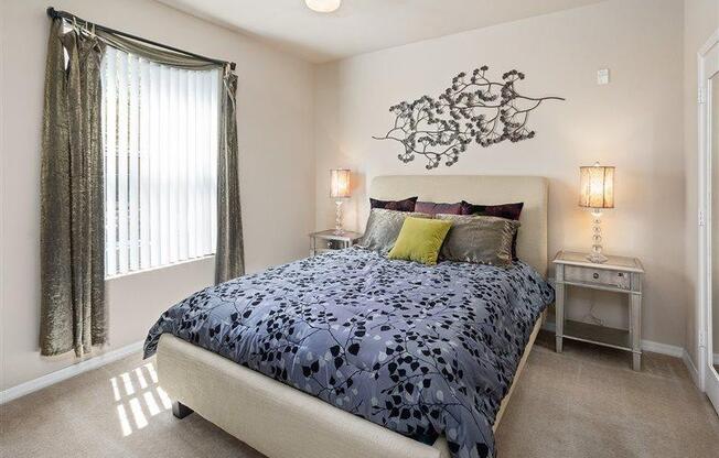 Photo of a model apartment bedroom with queen size bed, blue bed spread and gray pillows. Large window lets sunlight in while two end tables with small lamps provide extra light