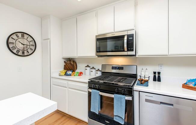 Modern kitchen featuring white cabinetry, stainless steel appliances, and a gas stove with blue towels hanging. A microwave is mounted above the stove. Thereâs a wall clock, utensils, and some food items on the countertop next to a sink. The floor is wood, and the overall design is bright and airy.