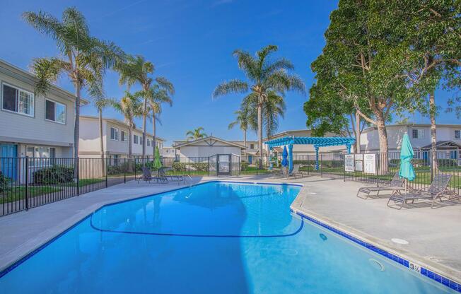 A sunny outdoor pool area surrounded by palm trees, with lounge chairs and umbrellas. The pool is clear blue, and nearby are residential buildings with a vibrant green landscape. A shaded seating area is visible under a blue pergola. The scene conveys a serene and inviting atmosphere.