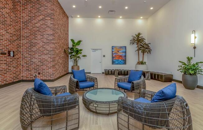 A modern indoor lounge area featuring four woven chairs with blue cushions arranged around a round glass table. The space has a brick wall, potted plants, and a piece of abstract art. Soft lighting enhances the inviting atmosphere, creating a cozy gathering spot.