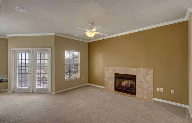 A spacious and inviting living room here at Mission Green featuring warm neutral walls, plush carpeted flooring, a tiled fireplace as a focal point, a ceiling fan with overhead light, and large windows with blinds alongside glass patio doors that allow natural light to brighten the open and comfortable space.