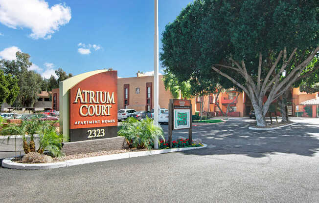 A striking community monument sign here at Aspire Pinnacle Park featuring bold lettering, the address number displayed prominently, and landscaped surroundings with palm trees and colorful flowers. A shaded drive leads into the property with mature trees and warm Southwestern-style buildings creating a welcoming and well-maintained entrance experience.