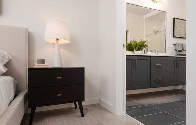 a bedroom with a black nightstand with a lamp and a bathroom with a sink