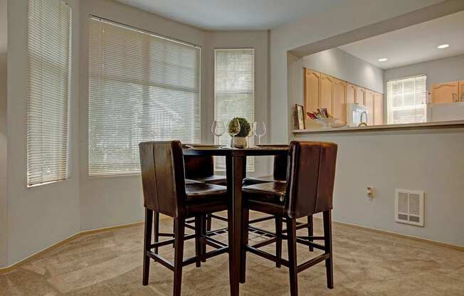 A cozy dining nook here at Aspen Creek featuring a tall square dining table with four upholstered chairs set on soft carpet, surrounded by multiple windows with blinds that bring in natural light, and positioned beside a breakfast bar opening into the kitchen with warm wood cabinetry and clean, neutral finishes.