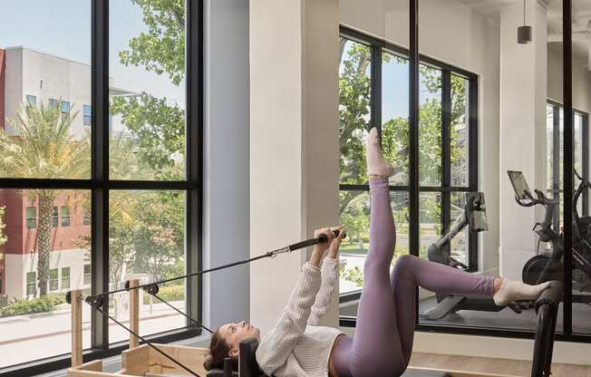 A woman in a purple outfit is doing a pilates exercise on a reformer in a spacious room with large windows.
