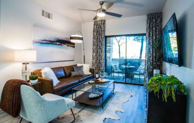 A living room with a brown leather couch, a grey armchair, a glass coffee table, and a flat-screen TV mounted on the wall.