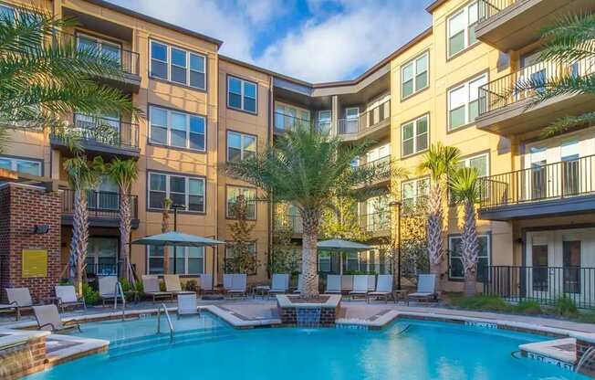 A swimming pool is surrounded by lounge chairs and palm trees in front of apartment buildings.