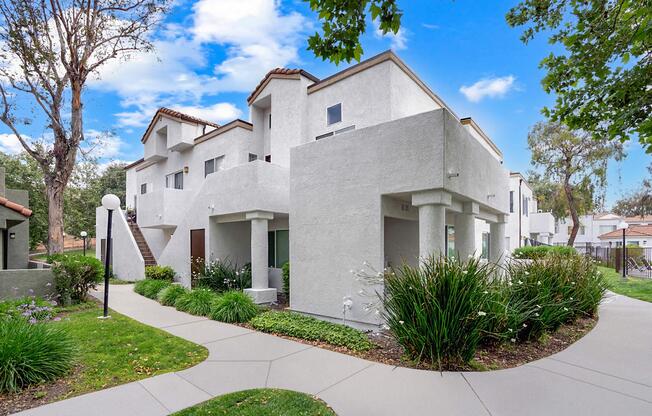 A modern, three-story white building with a stucco exterior, surrounded by greenery and well-maintained lawns. Pathways lead to the entrance, flanked by flower beds. Trees and blue skies are visible in the background, adding to a pleasant, residential atmosphere.