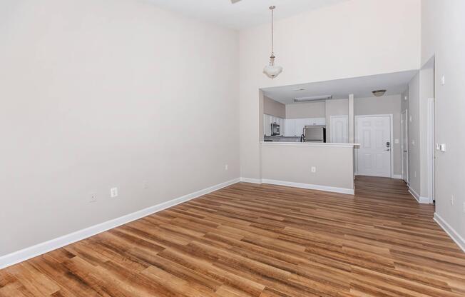 Spacious interior of an apartment with light-colored walls and wooden flooring. The area features a ceiling fan, a kitchen visible in the background, and a front door. Natural light fills the room, creating a bright and open atmosphere, ideal for decorating and personalizing.