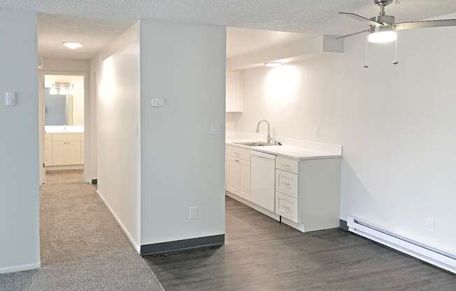 Apartment with a hallway on the left and kitchen on the right with faux wood flooring, white cabinets and appliances and a ceiling fan.