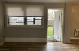 A room with a window covered by white blinds and a door open to a backyard.