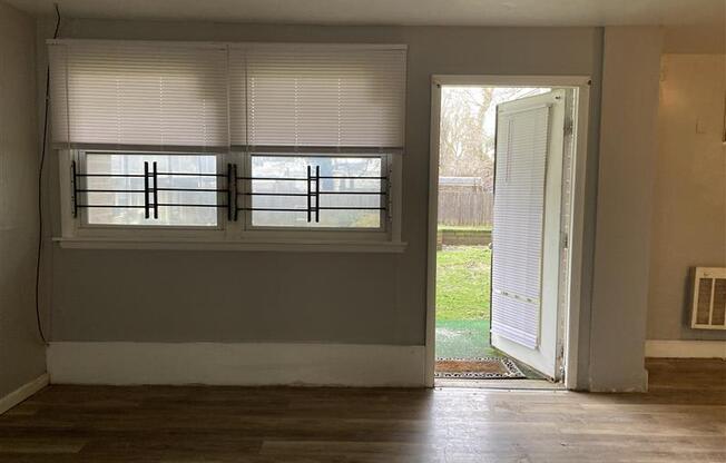 A room with a window covered by white blinds and a door open to a backyard.