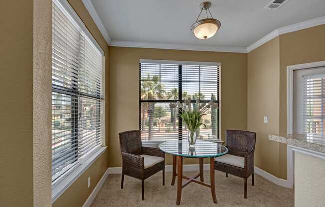 A bright dining nook here at Tuscany at Faudree with plush carpet and neutral walls. A small round glass-top table with two chairs sits beneath a ceiling light, while large windows with blinds bring in natural light and views of surrounding greenery, creating a cozy space for everyday meals.