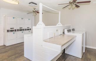 A laundry room featuring several washing machines and dryers neatly arranged against the wall, with countertops for folding laundry. The space is well-lit with ceiling fans and a clean, modern design, highlighted by a light color scheme and wooden flooring.