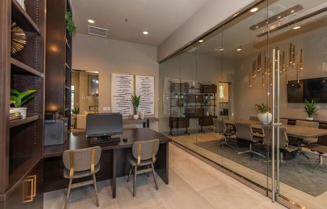 A modern office space featuring a workspace with a desk and two chairs, alongside a glass-walled conference room equipped with a large table and several chairs. The decor includes plants and decorative lighting, creating a professional and inviting atmosphere.