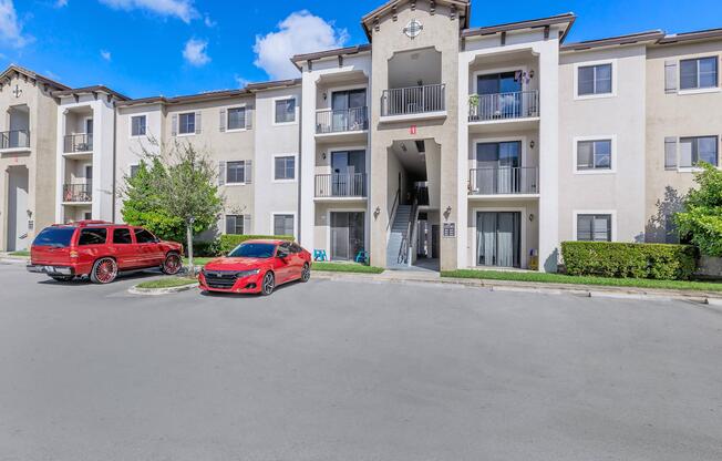 A residential apartment community featuring three floors with balconies, surrounded by greenery. Two cars, one red and one grey, are parked in the lot. The building has a light exterior with decorative elements on the top and large windows on each floor. Blue sky and clouds are visible above.