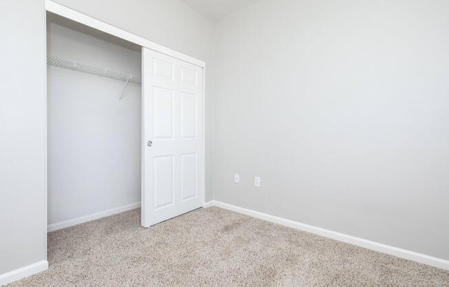 A clean, minimalistic room with light gray walls and beige carpet. A closet with a white door is visible, partially open, revealing a wire shelf inside. The room is empty, creating a spacious feel with no furniture or decorations.