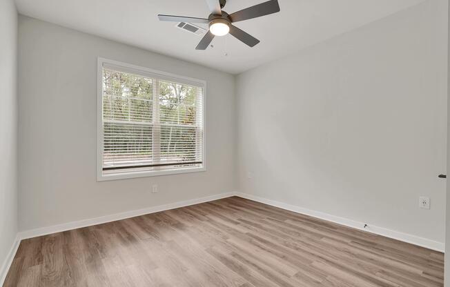 A minimalist room with light gray walls and a ceiling fan. The floor is covered with light wood laminate. A large window with white blinds allows natural light to enter, showcasing a view of greenery outside. The space is empty and ready for furnishing.