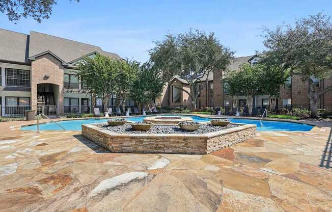 A pool surrounded by a stone patio with a fountain in the middle.