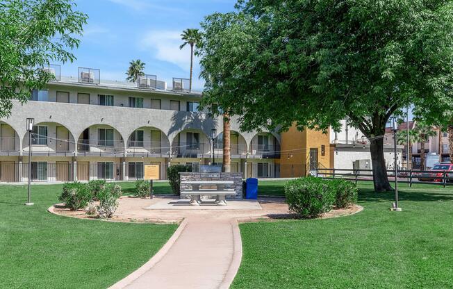 A landscaped park area featuring a paved walkway leading to a bench. Surrounding the bench are small shrubs and grass, with palm trees in the background. An apartment building with arched windows is visible, alongside a blue trash bin. The scene is bright and sunny, emphasizing a tranquil outdoor space.