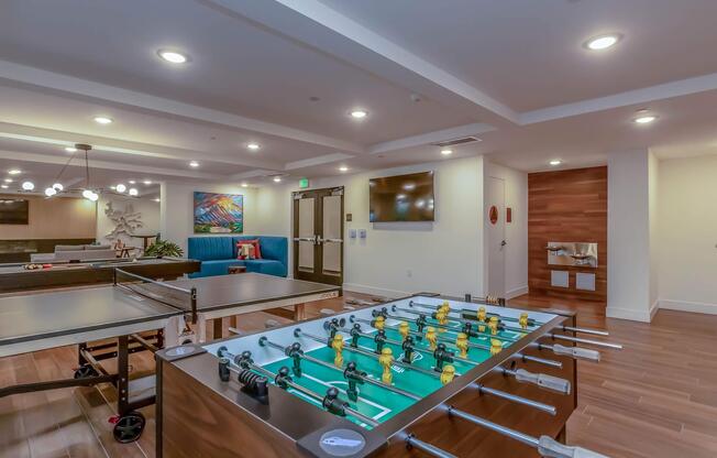 A recreational area featuring a foosball table and a ping pong table, with modern furnishings and decor. The space has bright lighting, a large wall-mounted TV, and comfortable seating. A colorful artwork piece is displayed on the wall, creating a vibrant atmosphere for socializing and entertainment.